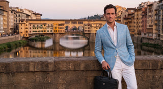 Man in a light blue blazer and white pants standing by a waterfront with buildings in the background
