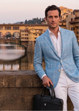 Man in a light blue blazer and white pants standing by a waterfront with buildings in the background